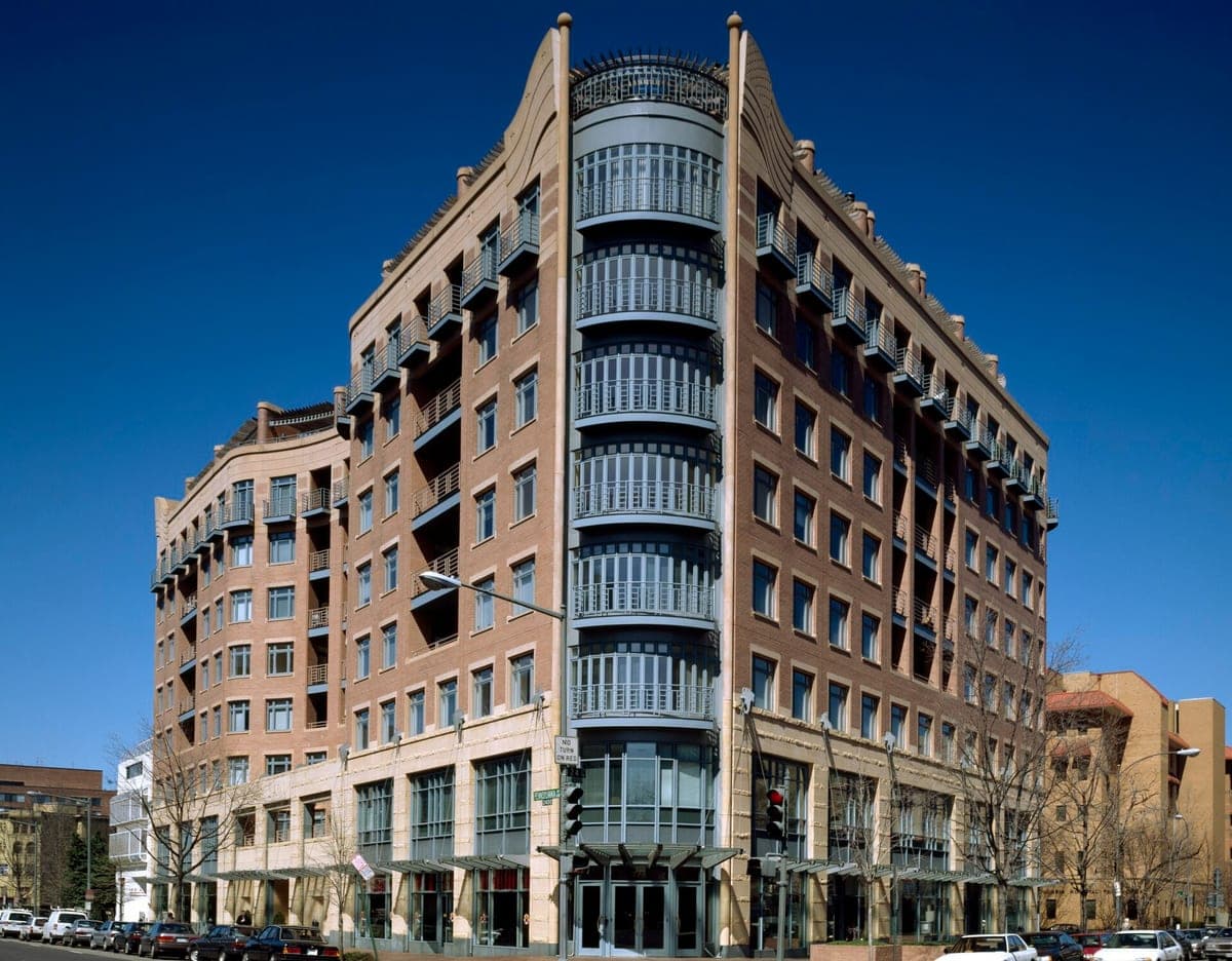 A photo of a corner office building in Washington, D.C., in tan and red brick, with a round glass tower of balconies at the corner, under a deep blue sky.