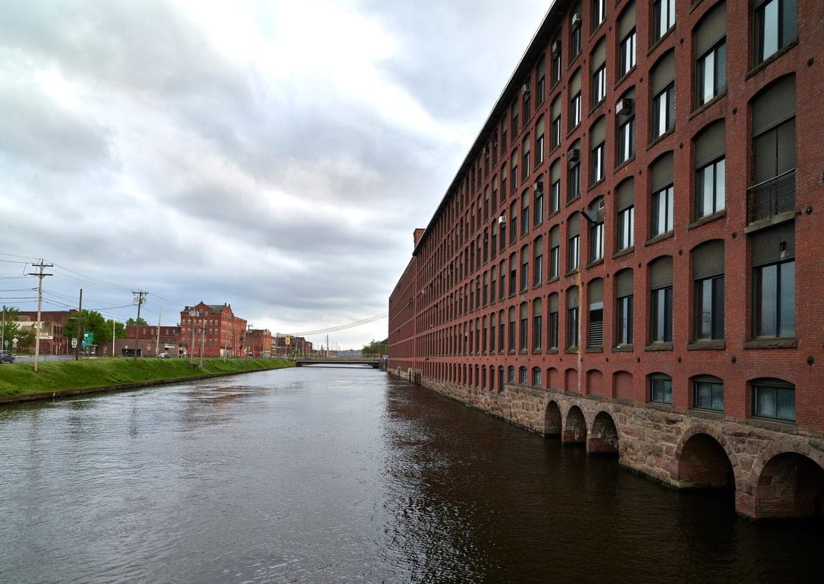 A photo along a canal of a long red brick mill in Holyoke, Massachusetts, with rows of tall windows above stone arches at the waterline.