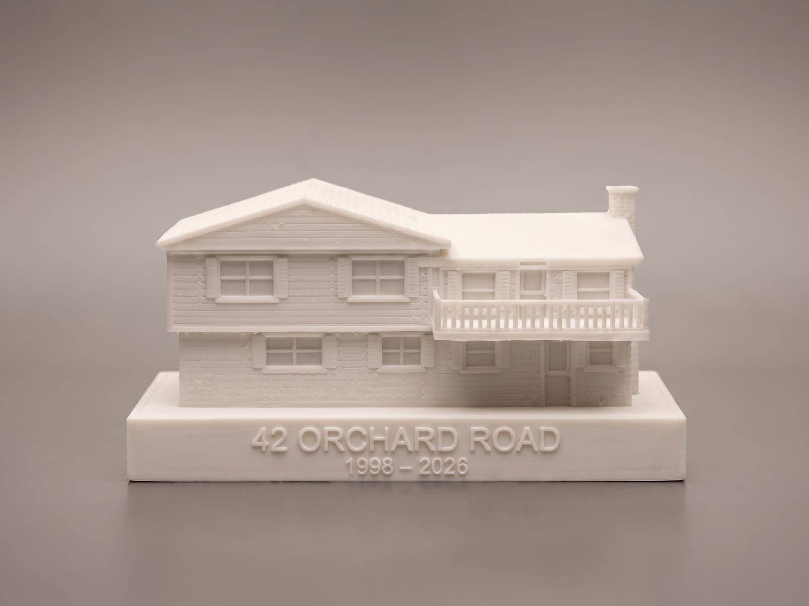 A finished split-level house piece with a railed deck on the upper floor, pictured on a slim base in the same white with raised letters reading 42 Orchard Road, 1998 to 2026, against a plain grey backdrop, in a generated picture