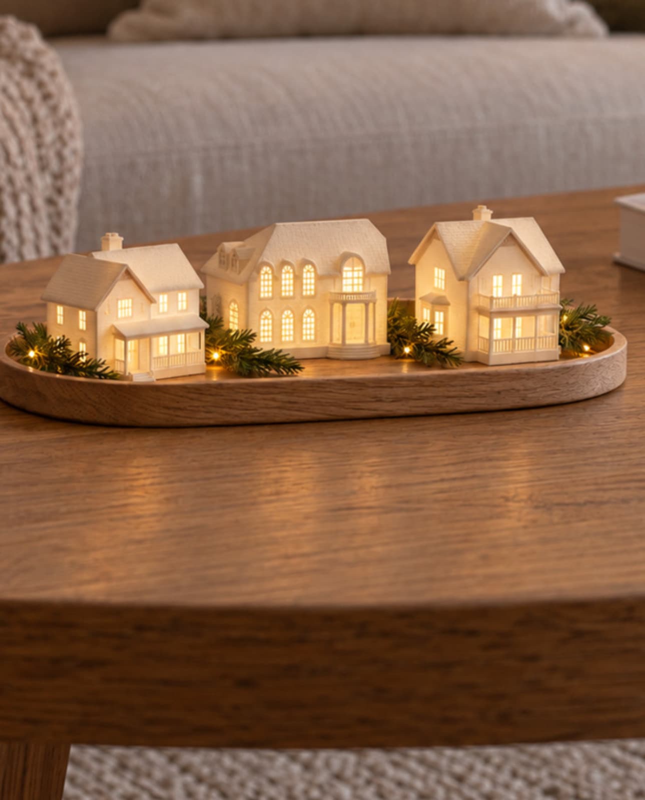 Three white house sculptures glowing from inside on a wooden tray with greenery on a living room coffee table