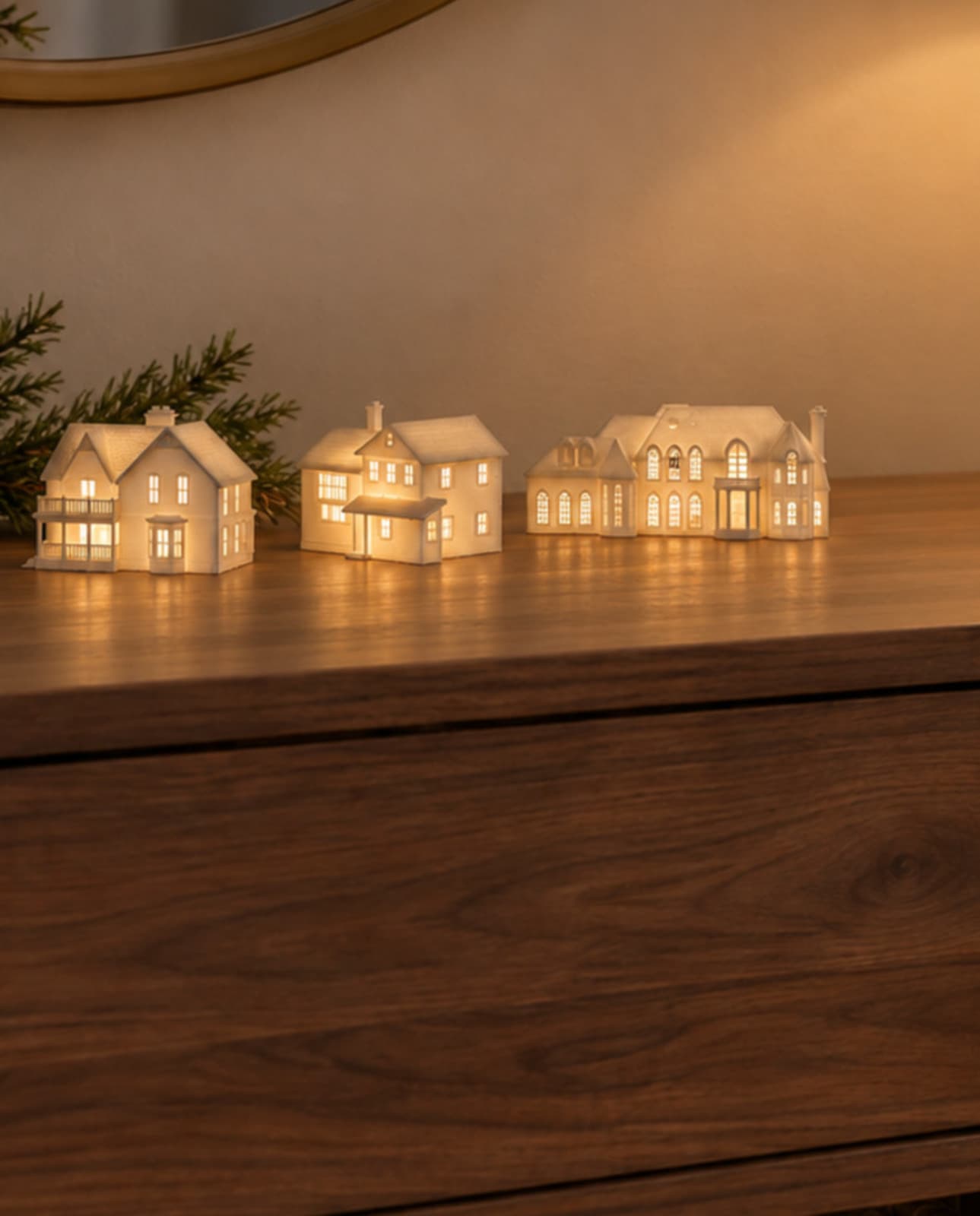 Three white house sculptures glowing from inside on a wood sideboard beside pine sprigs