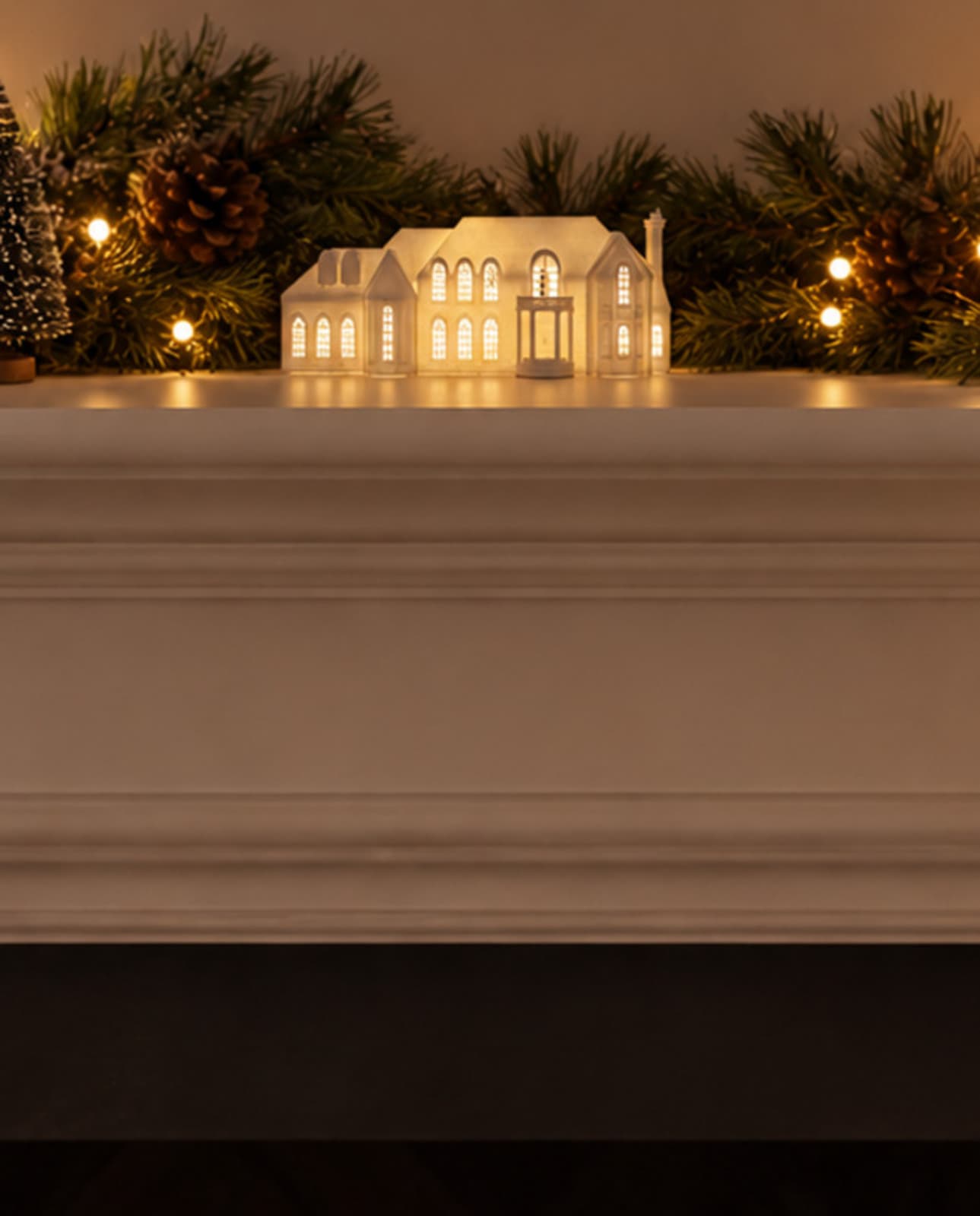 One white house sculpture glowing from inside on a white mantel among pine garland, pinecones and lights